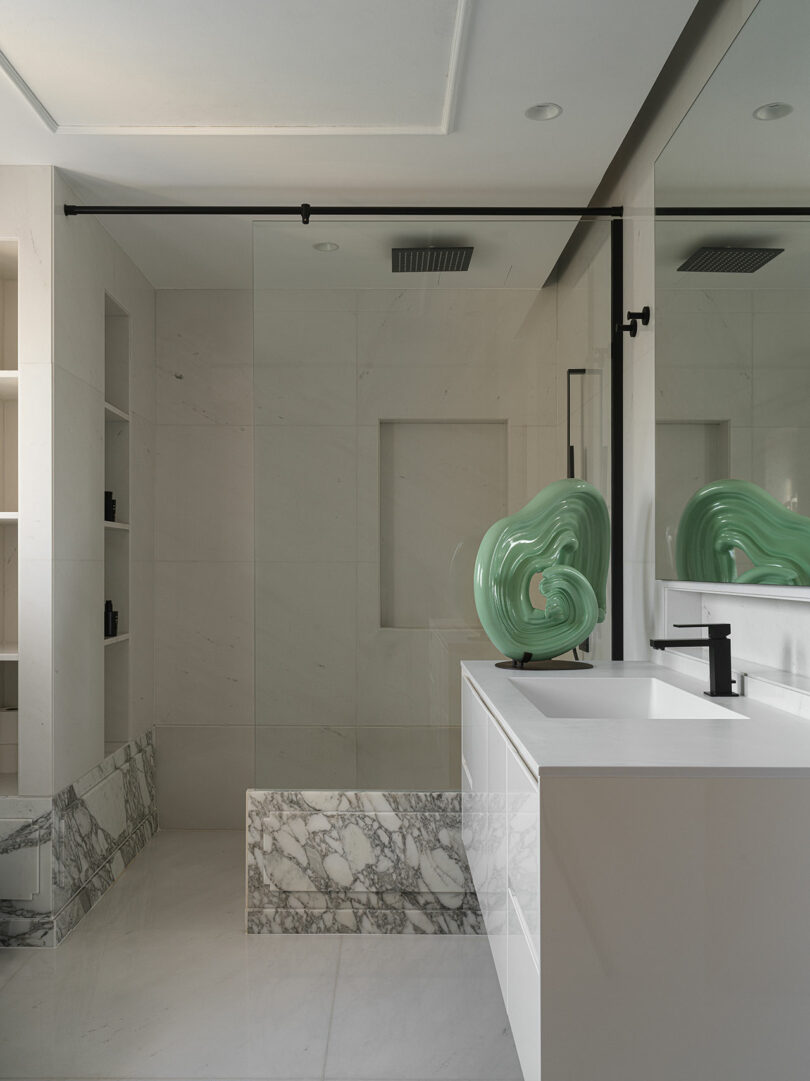 Modern bathroom with marble accents, a large frameless glass shower, sleek white cabinetry, and a green sculptural element on the counter, reminiscent of Marina Baisel's refined designs.