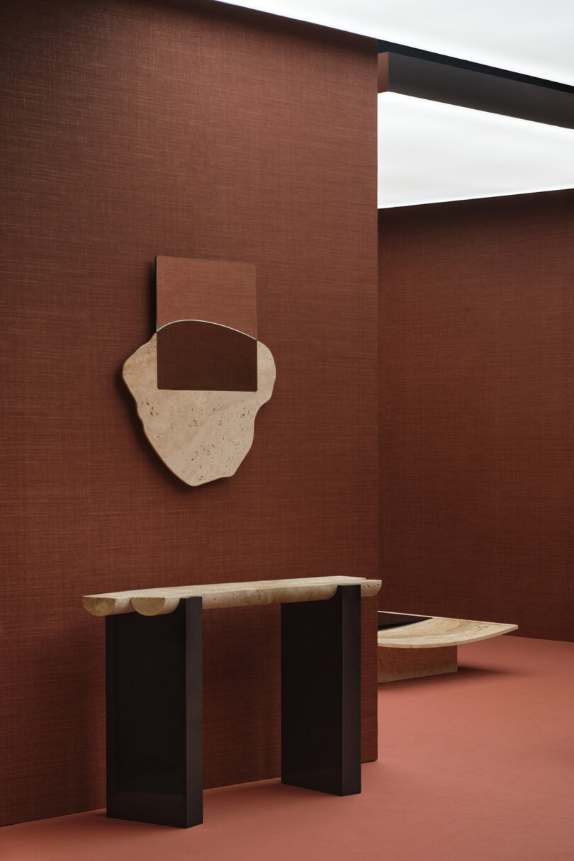 Modern interior with a stone console table and abstract wall mirror against a textured reddish-brown wall.