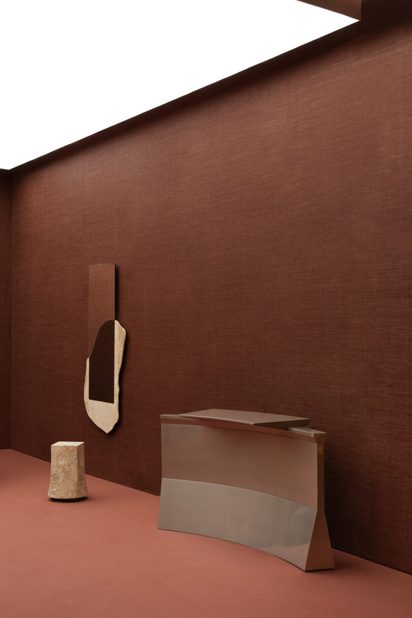 A minimalist room with brown walls and floor, featuring a geometric metal console, a rough-hewn stone stool, and an abstract wall art piece.