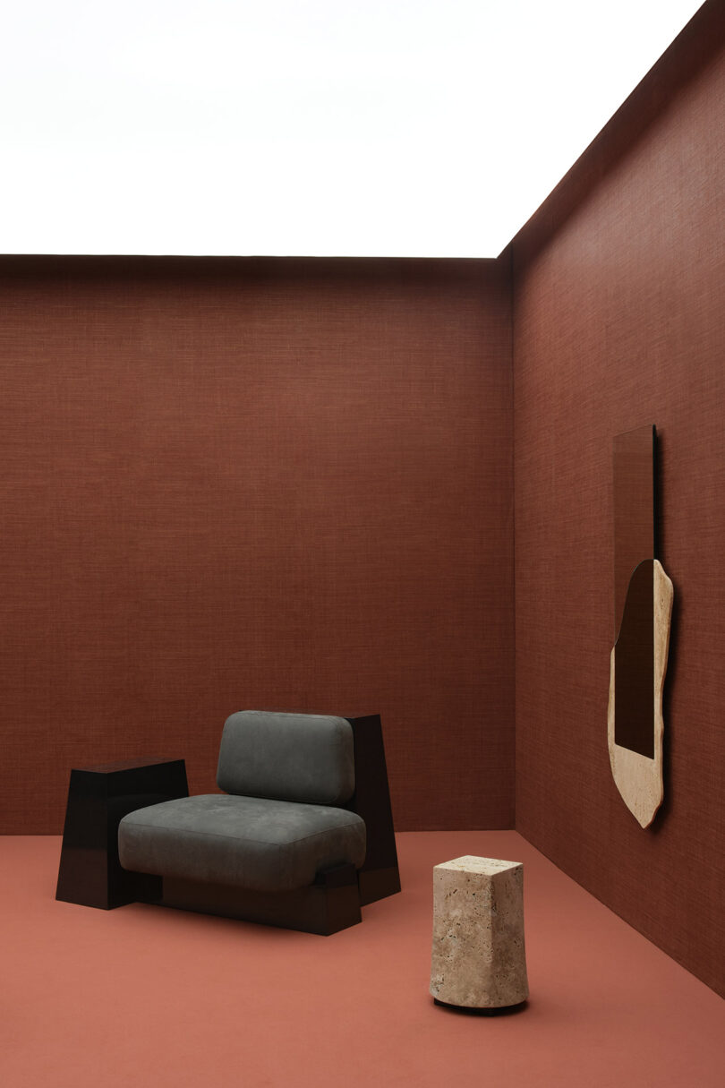 Minimalist room with brown walls and floor, featuring a gray armchair, a black side table, a round stone stool, and an abstract wall mirror.