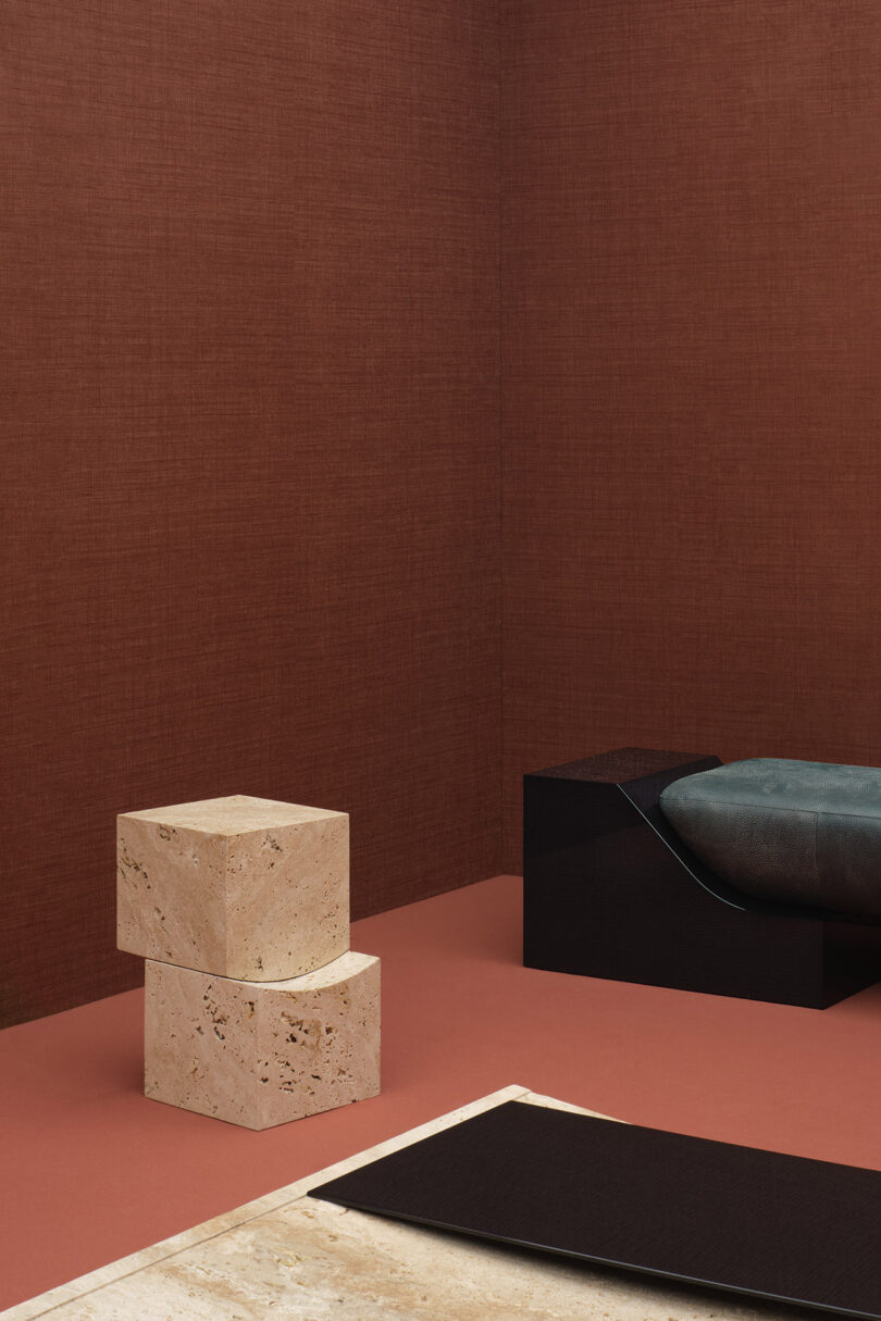 Minimalist room with brown textured walls, a black bench with a teal cushion, two stacked beige stone blocks, and a rectangular stone surface.