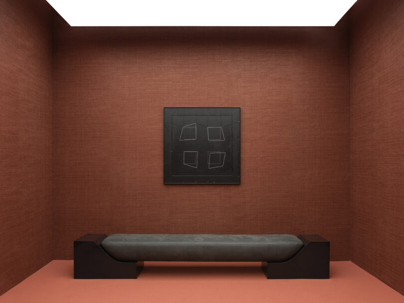 A minimalist room with brown walls features a gray bench and a black abstract painting with geometric shapes.