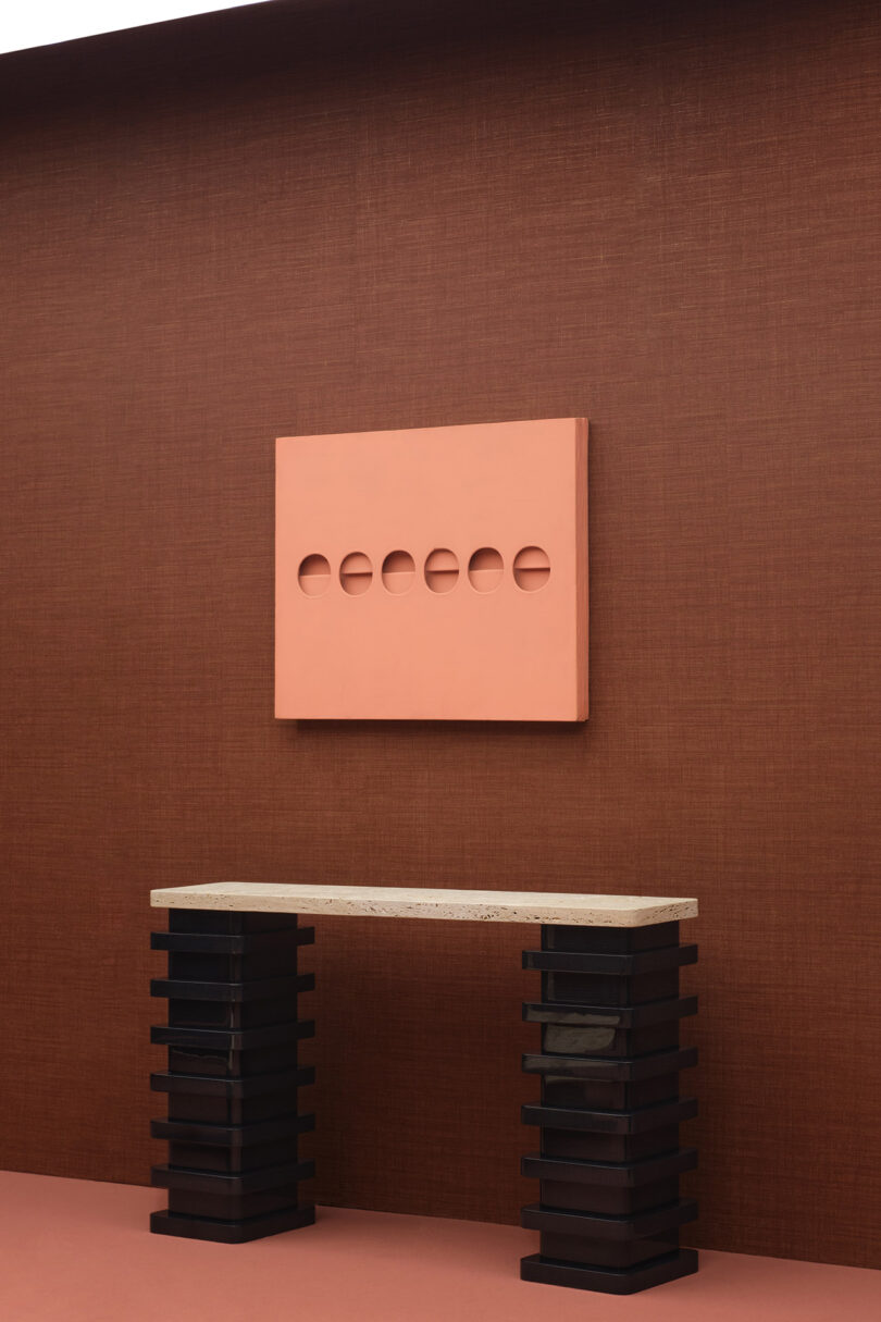 A minimalist setup with a geometric wall art, featuring six circular cutouts, hangs above a console table with a marble top and stacked black block legs, against a brown textured wall.