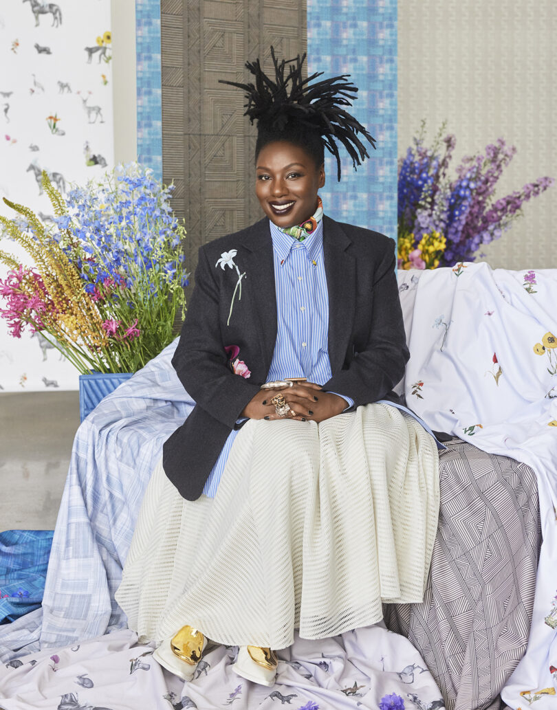 A person sitting on a draped surface with floral patterns, wearing a blazer, striped shirt, and white skirt. Colorful flowers and patterned wallpaper are in the background.