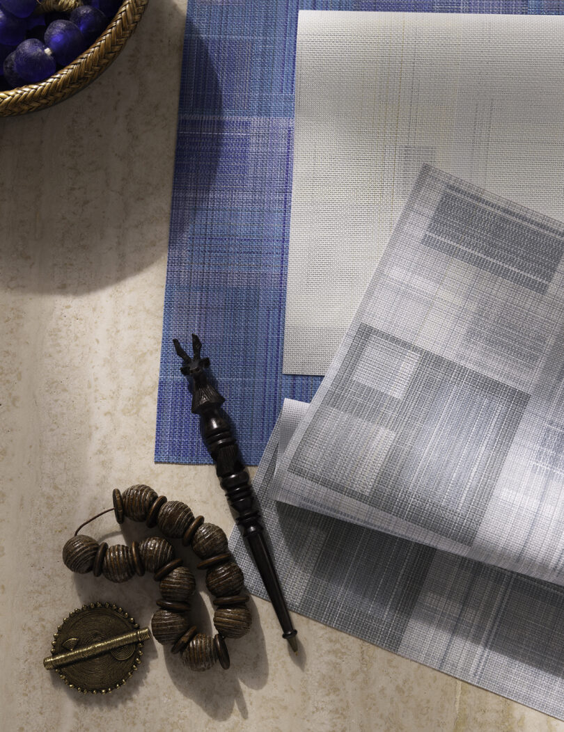 Beads and wooden pen are on a table with textured paper, a wicker basket with a beaded necklace inside.