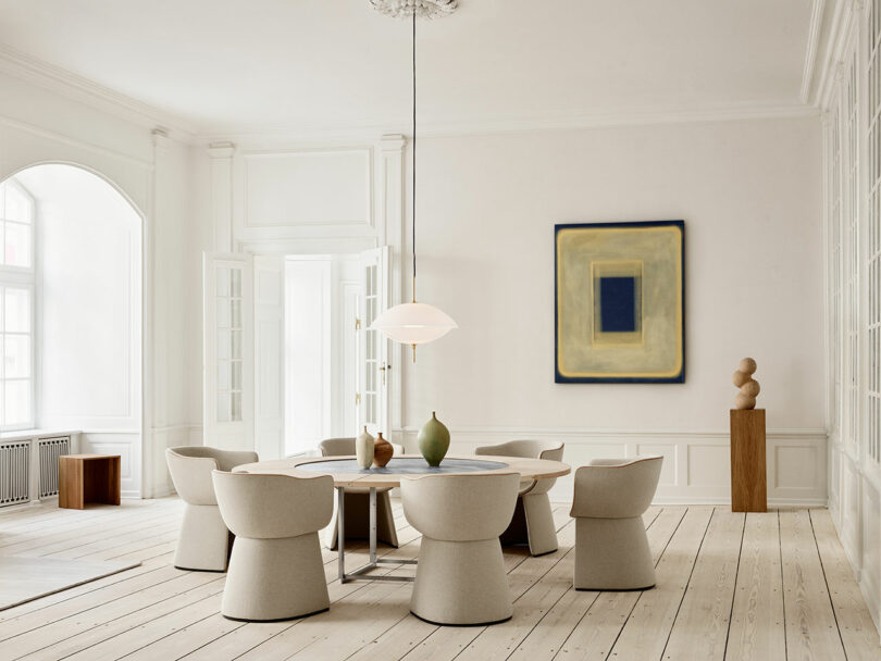 Elegant dining room with a circular table, six armchairs, a modern pendant lamp, large windows, and a geometric painting on the wall. Minimalist decor with natural light.