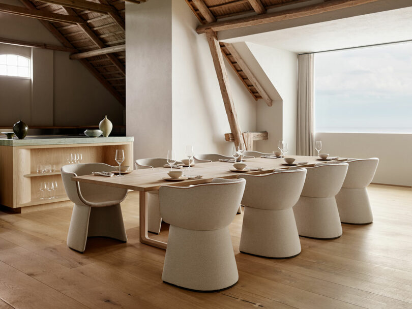 A modern dining room with a long table set for eight, featuring beige upholstered chairs. The room has wooden floors, exposed beams, and a large window offering a view of the sky and sea.