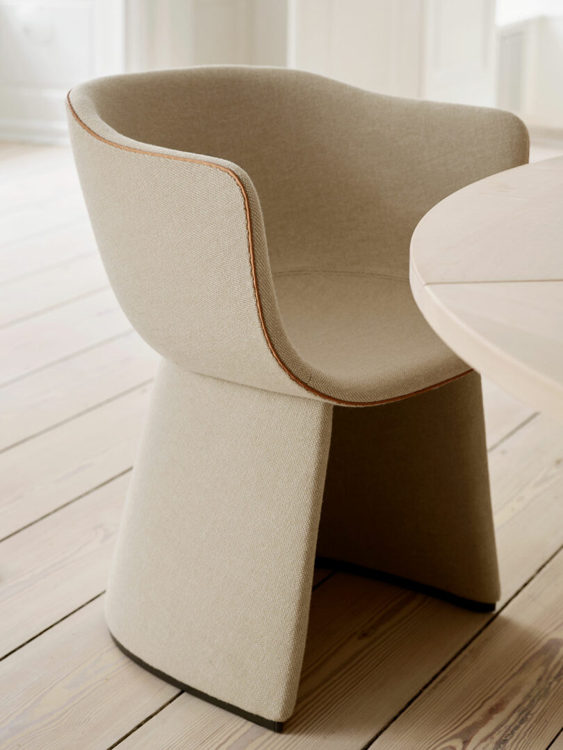 A beige upholstered chair with a curved back and seat, placed next to a round wooden table on a light wooden floor.