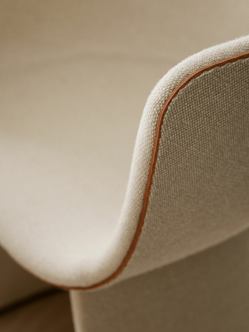 Close-up of a beige fabric chair with brown piping, showcasing the smooth curve of the armrest.