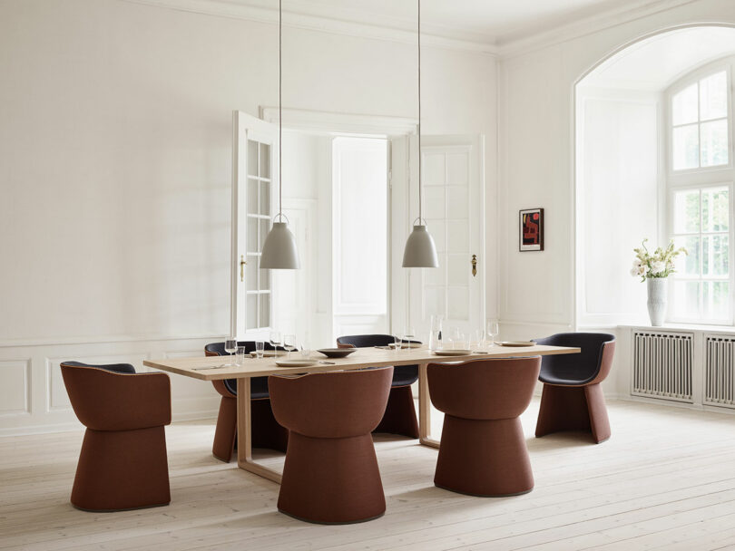 A modern dining room with a wooden table, six brown chairs, and two pendant lights. Large windows and white walls create a bright, airy atmosphere. A small artwork hangs on the wall.