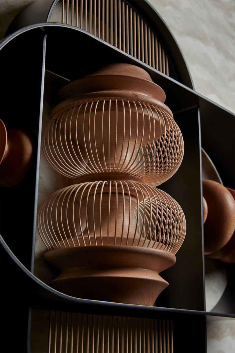 A close-up of modern wooden and wire sculptures displayed on a black metal shelf with circular compartments against a textured wall