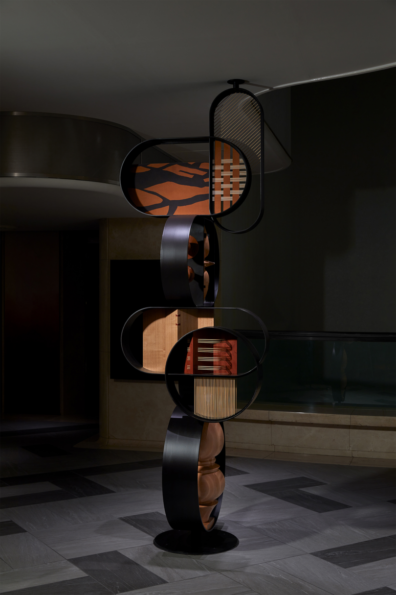 A modern abstract sculpture with circular and rectangular elements, featuring wood and metal materials, stands on a patterned floor in a dimly lit room