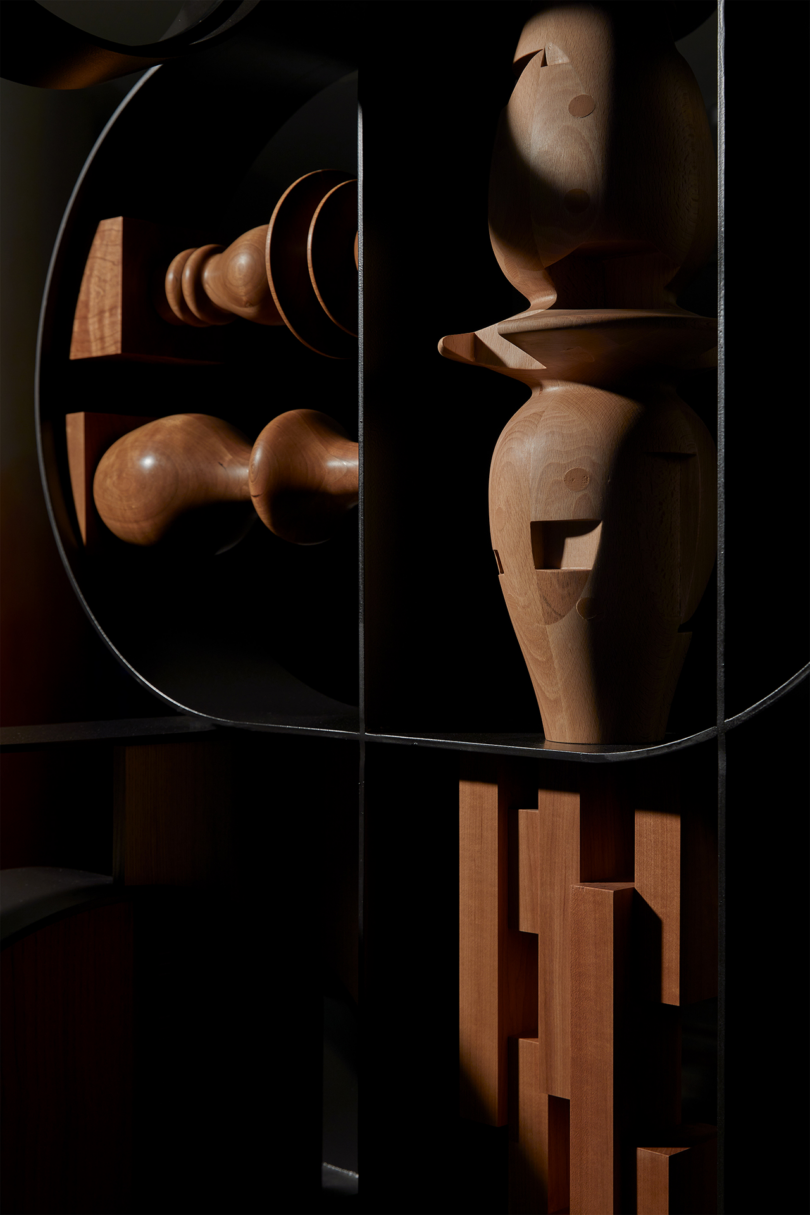 A wooden sculpture featuring various geometric shapes, including spheres, cylinders, and a segmented vase-like structure, illuminated with dramatic lighting