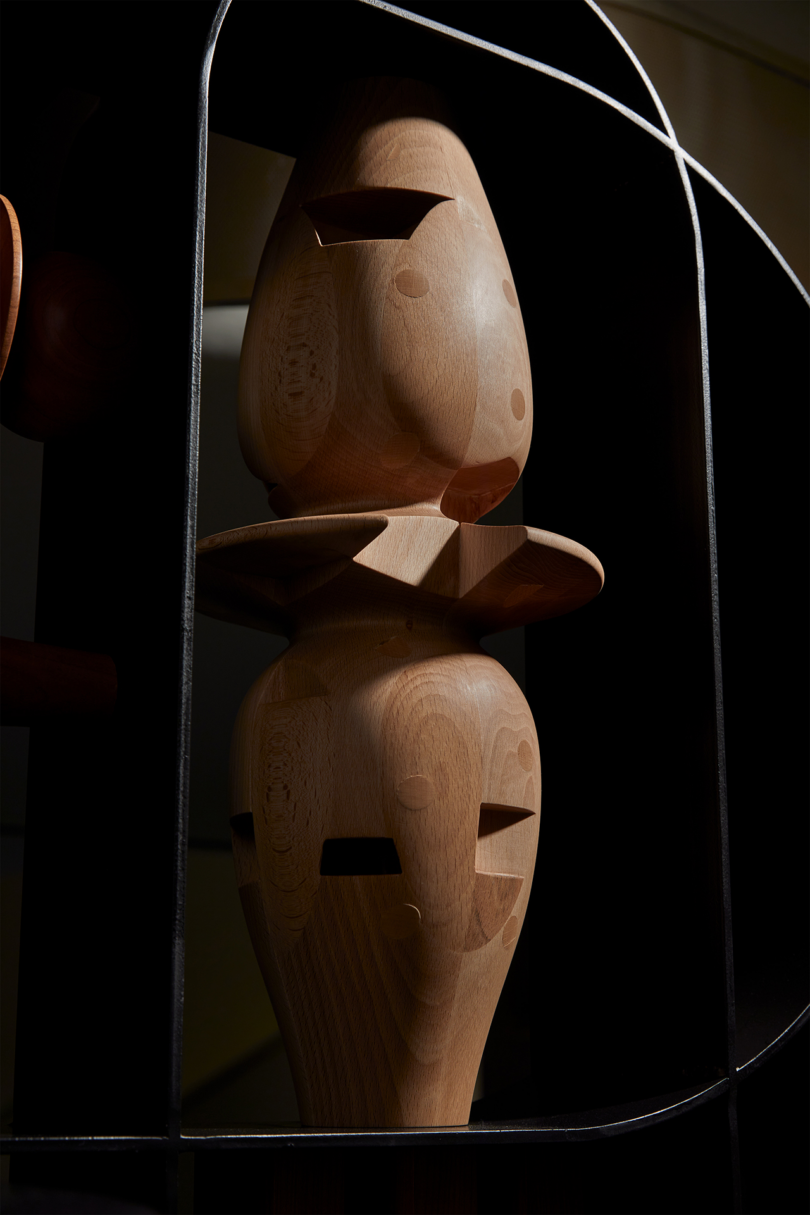 Two wooden figurines with minimalistic facial features are stacked one on top of the other inside a thin metal frame, casting shadows