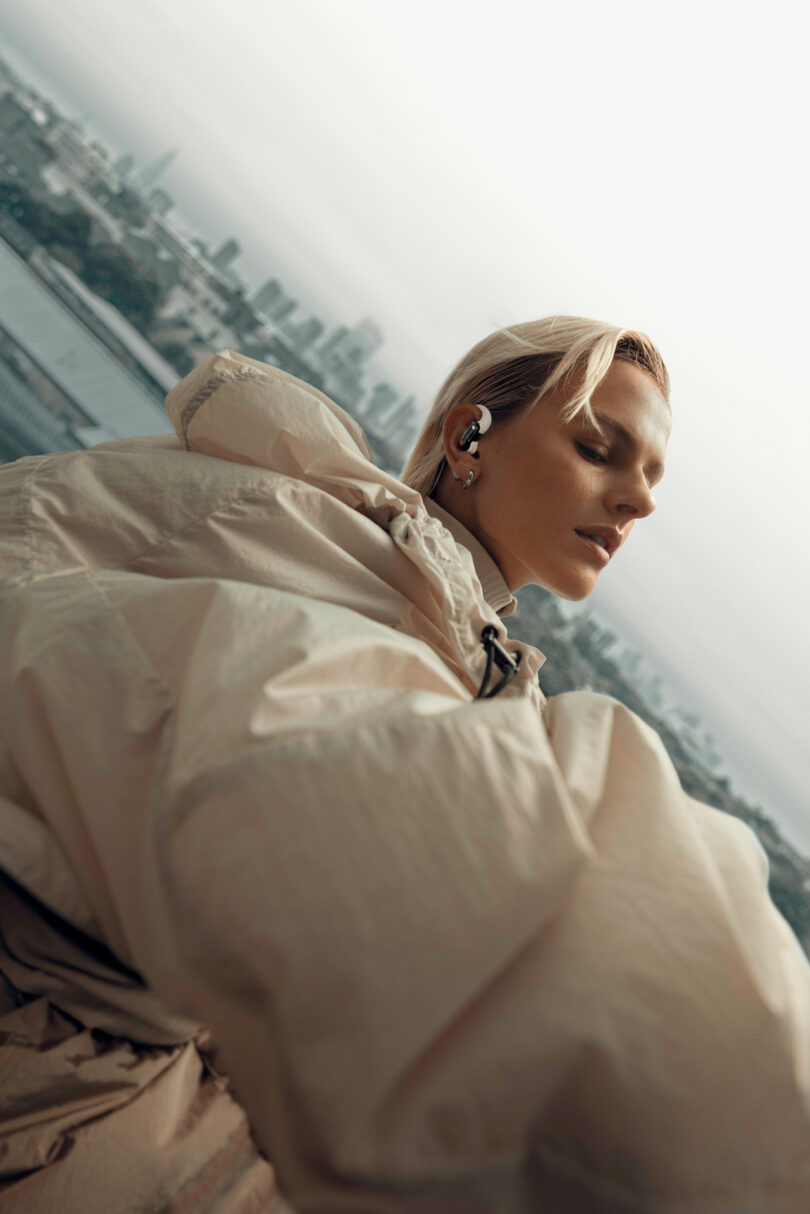 Person in a light-colored jacket wearing earbuds, standing outdoors with a cityscape in the background.