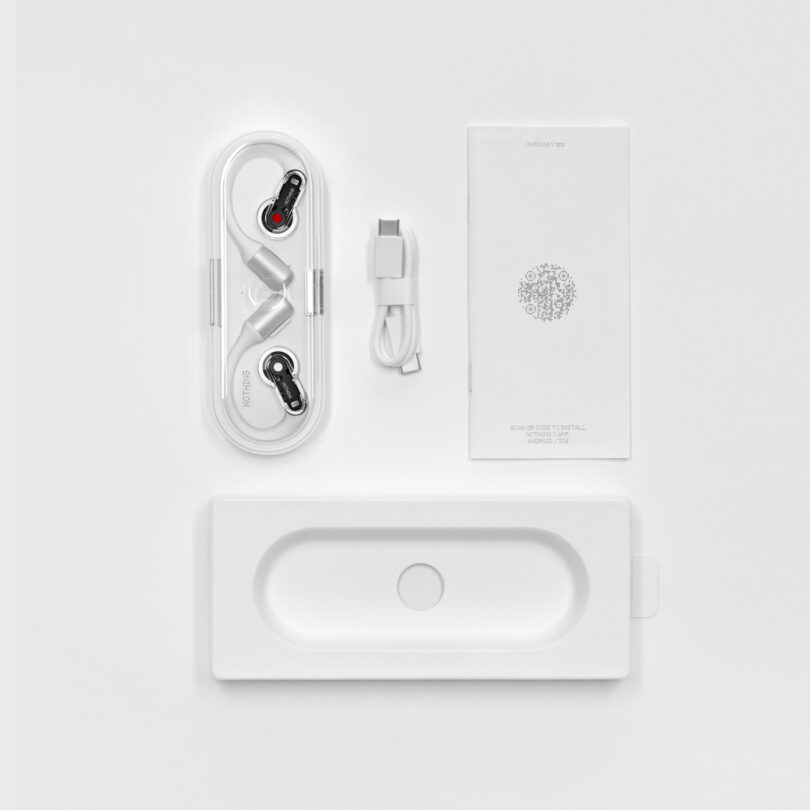 White earphones with attached wires in a plastic case, a USB-C cable, a small booklet, and an empty packaging tray all placed on a white surface.