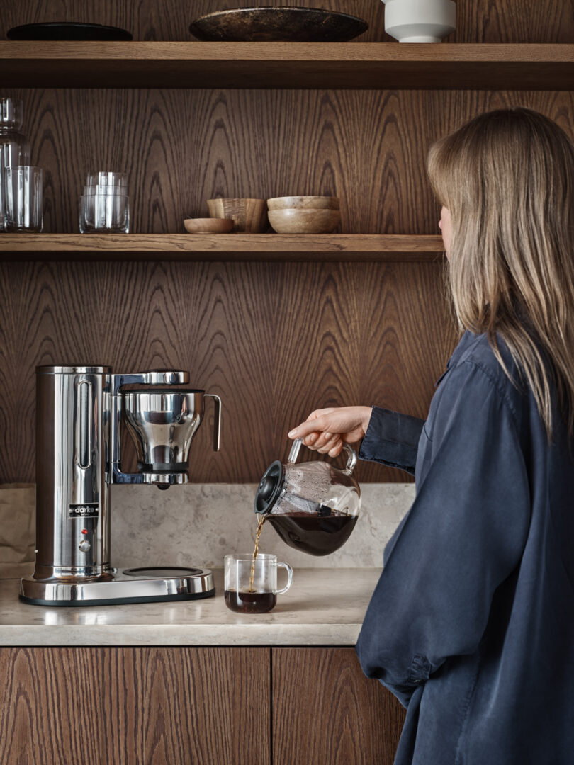 A person is pouring coffee from a glass carafe into a mug beside the sleek Aarke Coffee System on a kitchen counter with wooden shelves.