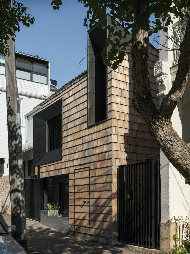 Three-story modern house with wooden shingle facade, large black-framed windows, and a tree in front. The house is flanked by a gated fence and neighboring buildings.