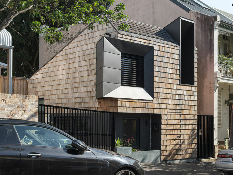 Modern house with shingle siding and large geometric window, fronted by a black fence and a parked car.