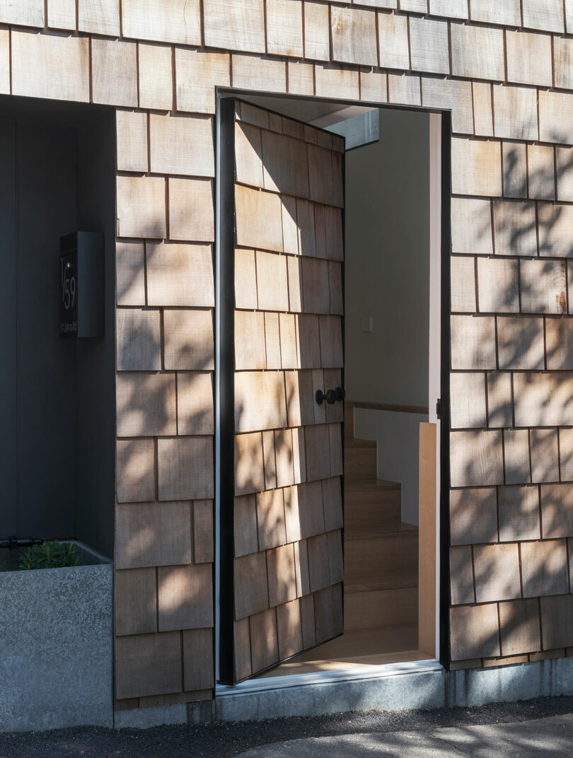 A wooden shingle wall with an open door leading to a staircase inside. The door casts shadows on the exterior.