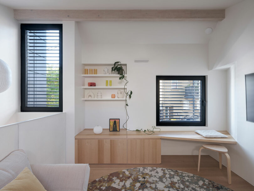 A minimalist room with a beige sofa, wooden bench, and windows. Shelves hold books and decor. A plant trails from a shelf, and a round rug is on the floor.