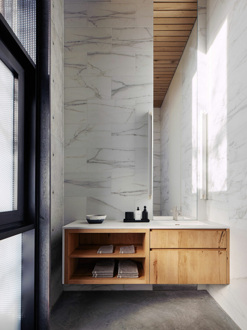 A modern bathroom features a wall-mounted wooden vanity with open shelves and cabinets, a rectangular sink, and a large mirror. The walls are lined with white marble tiles.