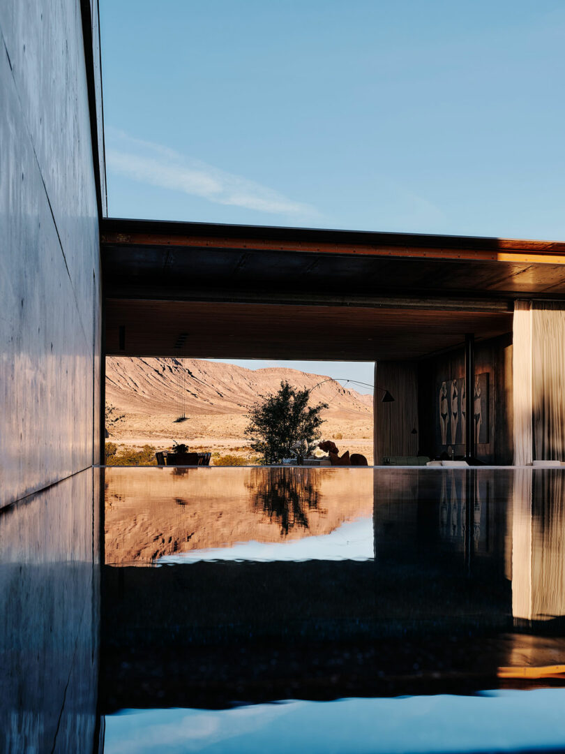 Modern house with open-air design featuring a reflective pool. In the background, desert landscape with rugged hills and clear blue sky.