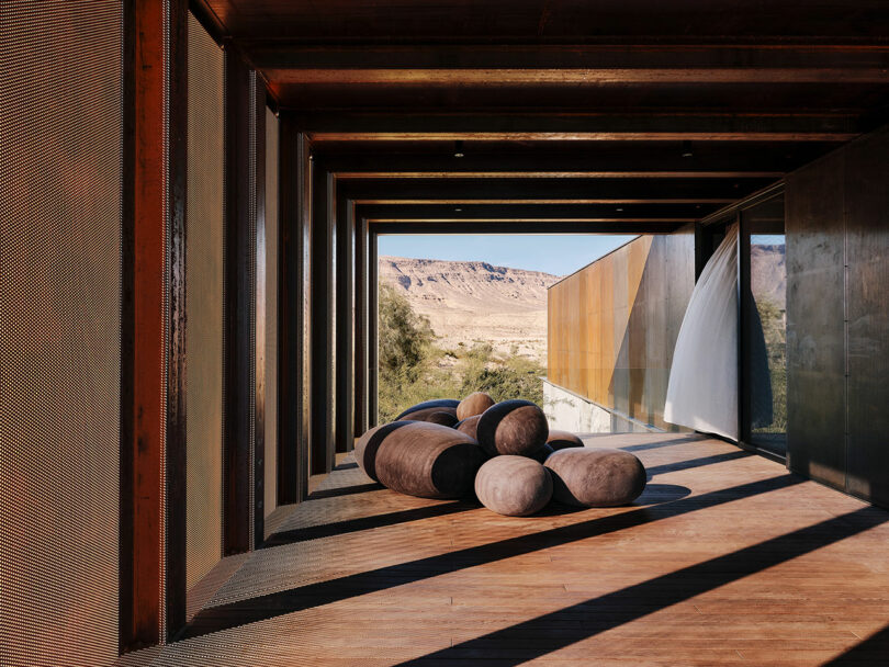 A modern outdoor walkway with wooden beams and mesh screens features an arrangement of cushion-like seating. Desert landscape is visible in the background.