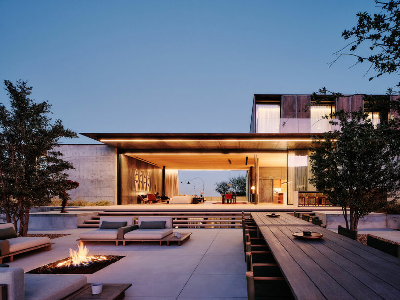 A modern, minimalist house with large glass walls, an open-plan living area, and an outdoor patio featuring a fire pit and wooden seating, set against a twilight sky.