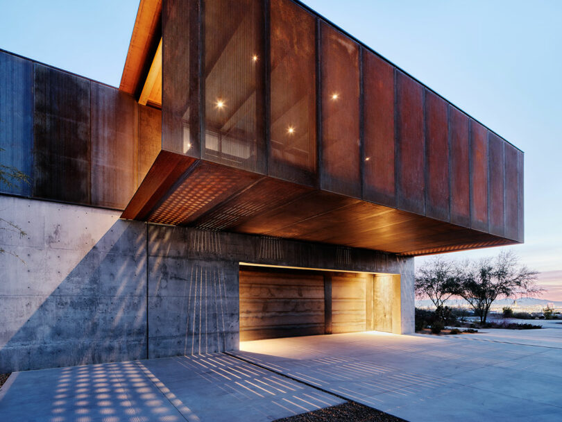 Modern building featuring an industrial design with concrete and rusted metal elements, including an overhanging second floor and large garage entrance. The structure is illuminated by outdoor lights.