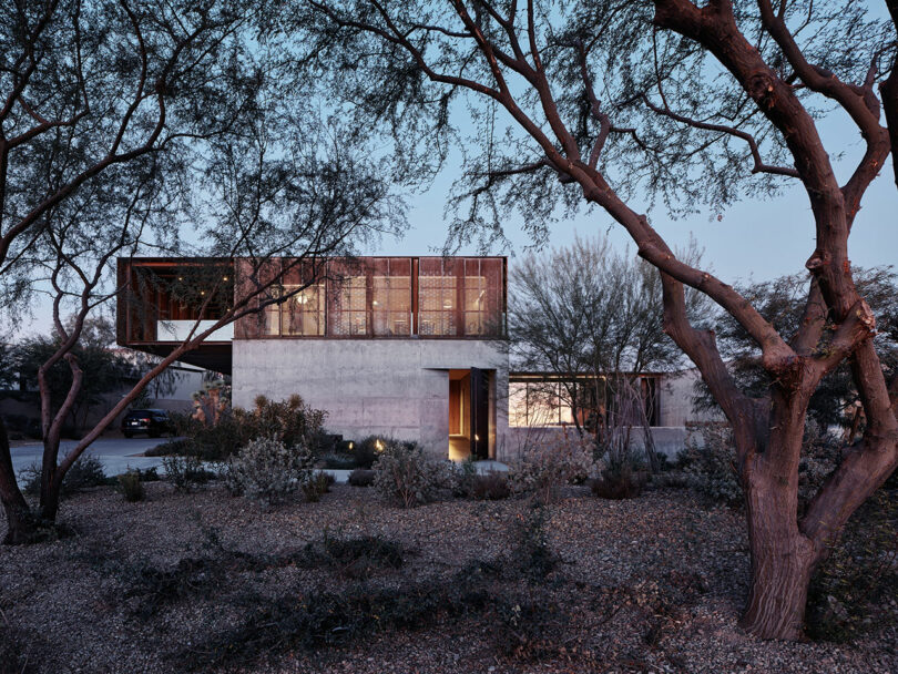 Modern two-story house with large windows and a minimalist design, set amidst a natural landscape with trees and shrubs.