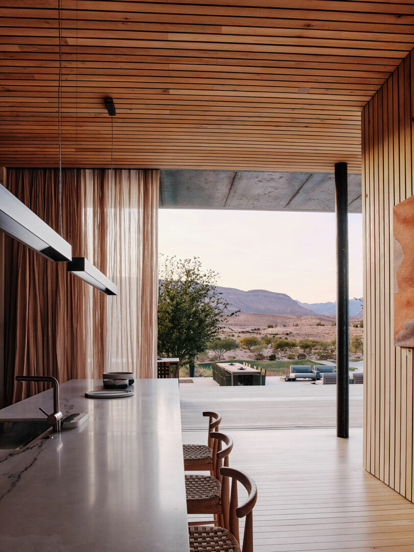 A modern kitchen with wooden ceiling and floor features a long counter, bar stools, and hanging lights. Large windows with sheer curtains offer a view of a desert landscape and mountains outside.