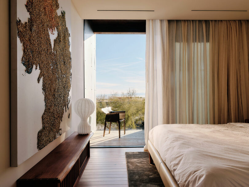 A bedroom featuring a large artwork, a white bed, a wooden bench, a white lamp, and a desk near a large window with sheer curtains, offering a view of an outdoor landscape.