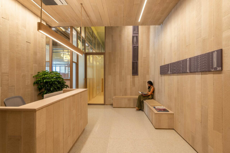 A modern reception area with wooden walls and ceiling, a reception desk, a plant, and a woman sitting on a bench reading a magazine.