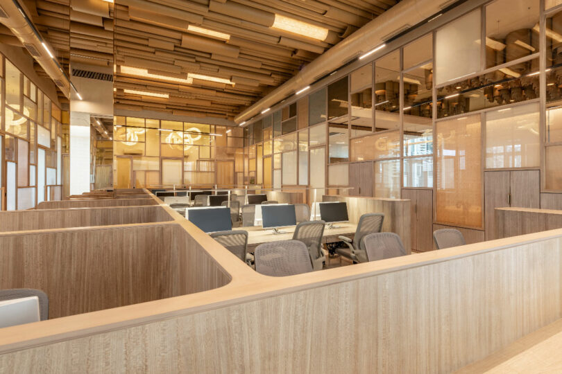 A modern office space with wooden partitioned workstations, computer monitors, ergonomic chairs, and large glass windows allowing natural light.