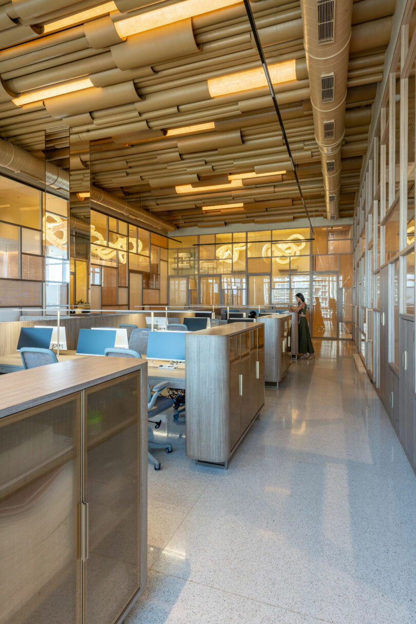 A modern office space with partitioned desks, blue and gray chairs, and wooden cabinetry. A single person is seen standing near the glass-walled conference room in the background.