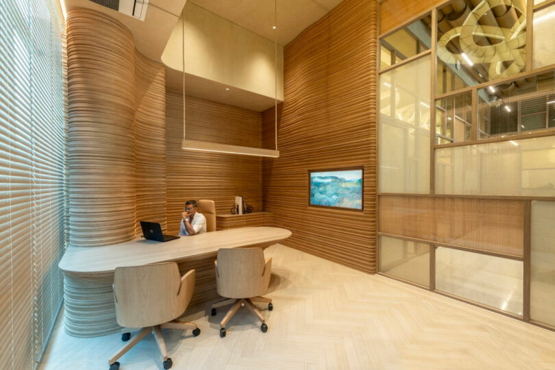 A person sits alone at a modern wooden desk in a sleek, contemporary office with curved wooden paneling, a wall-mounted screen, and large windows with blinds.