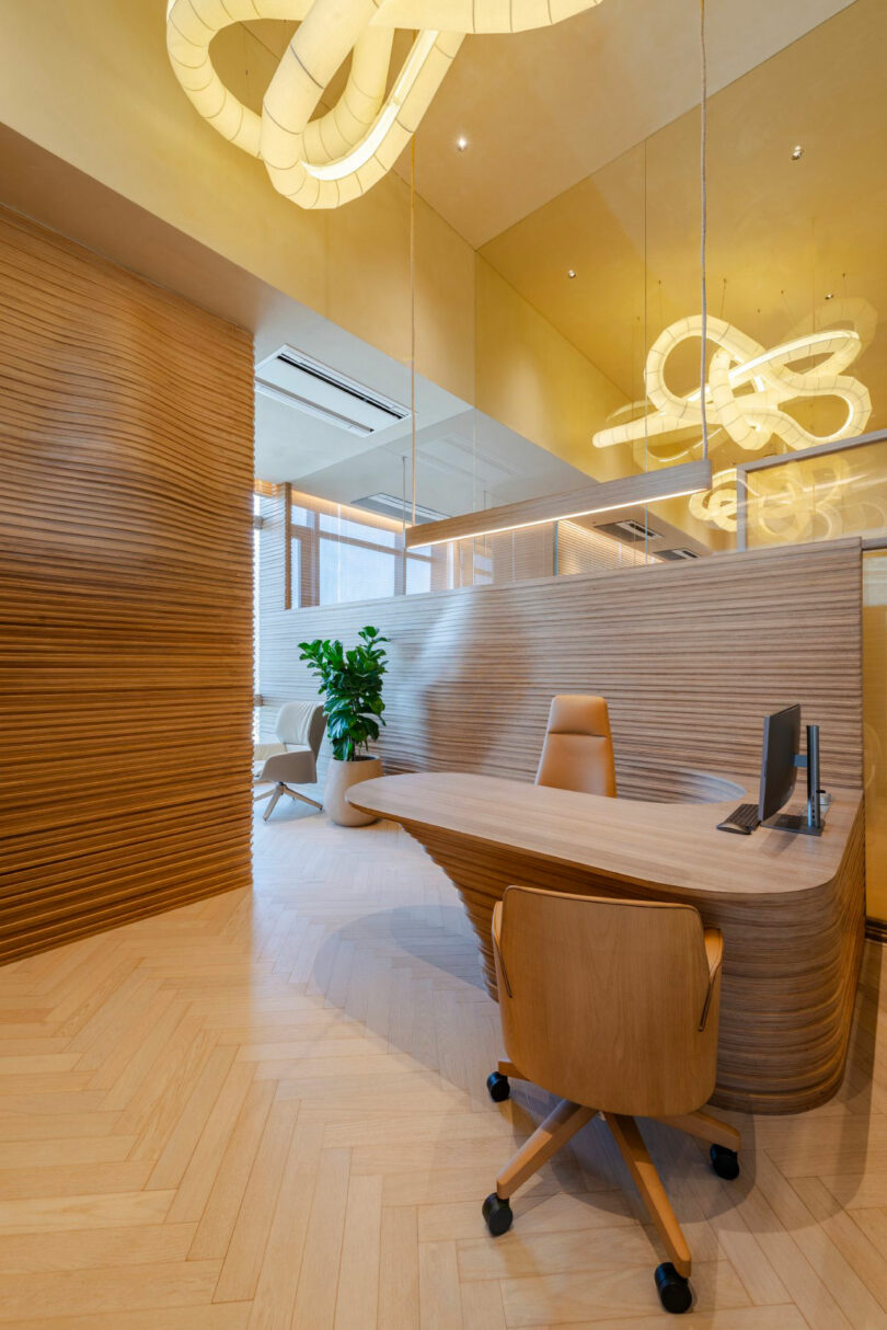 Modern office interior with wooden finishes, a unique geometric desk, an ergonomic chair, a computer monitor, a potted plant, and decorative lighting fixtures on the ceiling.