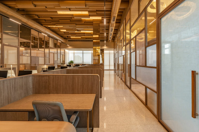 A modern office space with cubicles, wooden partitions, ergonomic chairs, and a mix of transparent and opaque paneling. The ceiling features geometric designs and the flooring is polished.