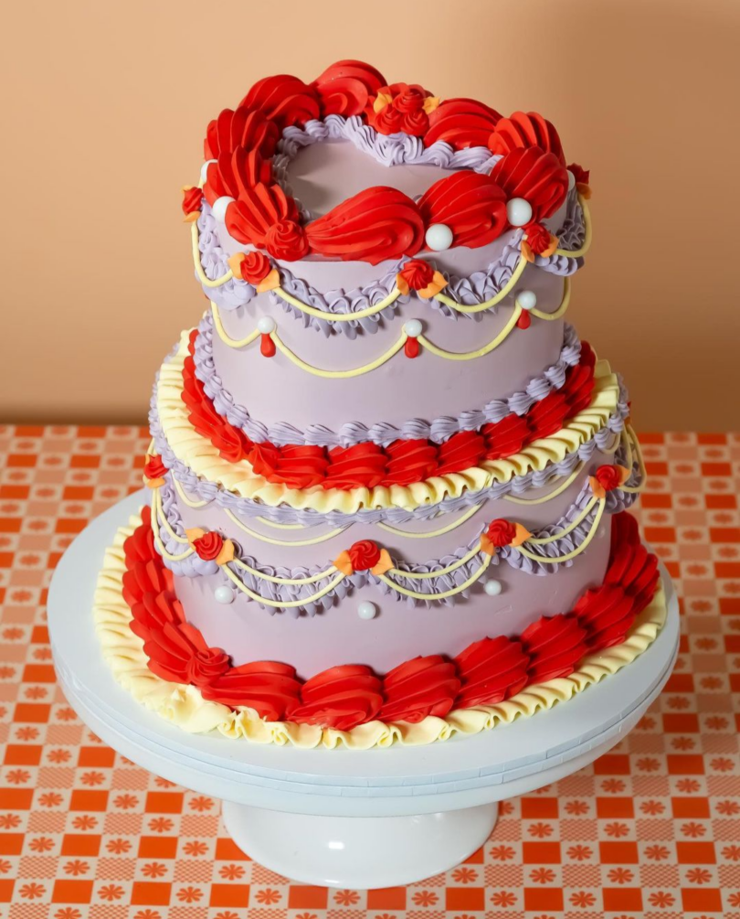 A three-tiered cake with light purple frosting, red and yellow decorative icing, and white pearls, displayed on a white cake stand with an orange checkered tablecloth background.