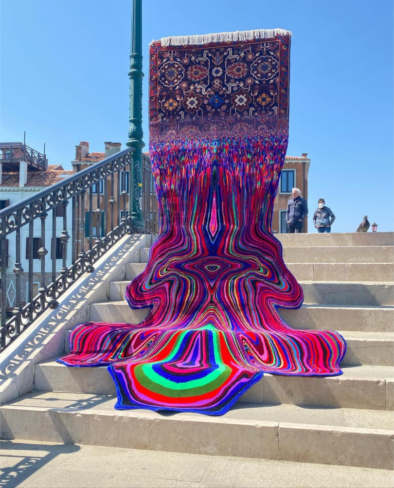 A traditional rug with intricate patterns appears to melt down a set of stairs in a surreal, distorted manner. Two people stand in the background near a handrail and historic buildings.