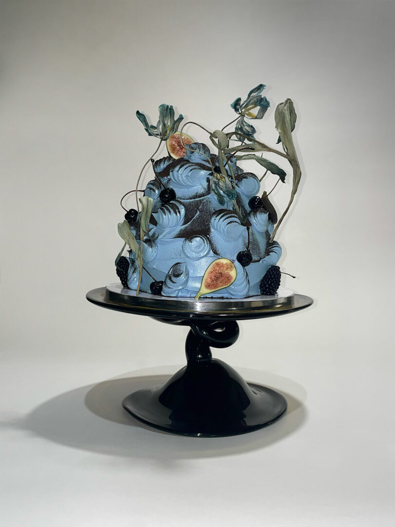 A decorative blue cake with figs, blackberries, and leafy accents sits on a black pedestal stand against a plain background.