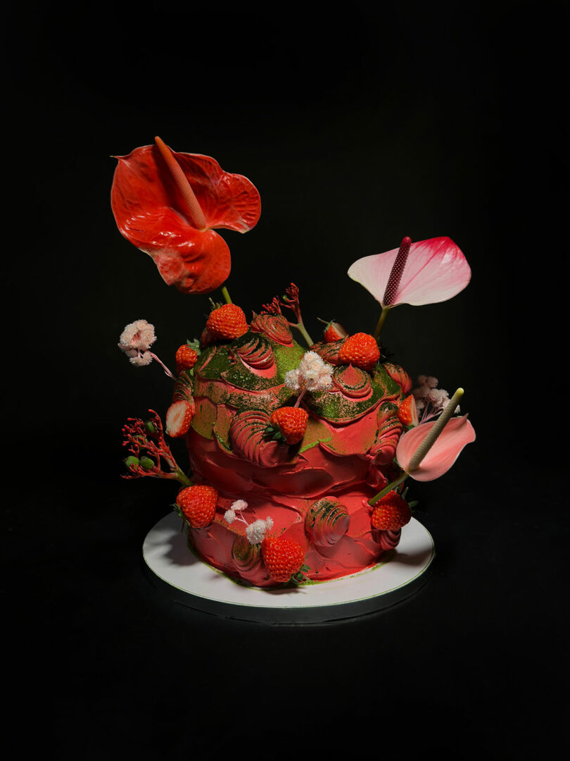 A two-tier cake with red and green frosting, topped and decorated with anthurium flowers, red berries, and small white blooms against a dark background.