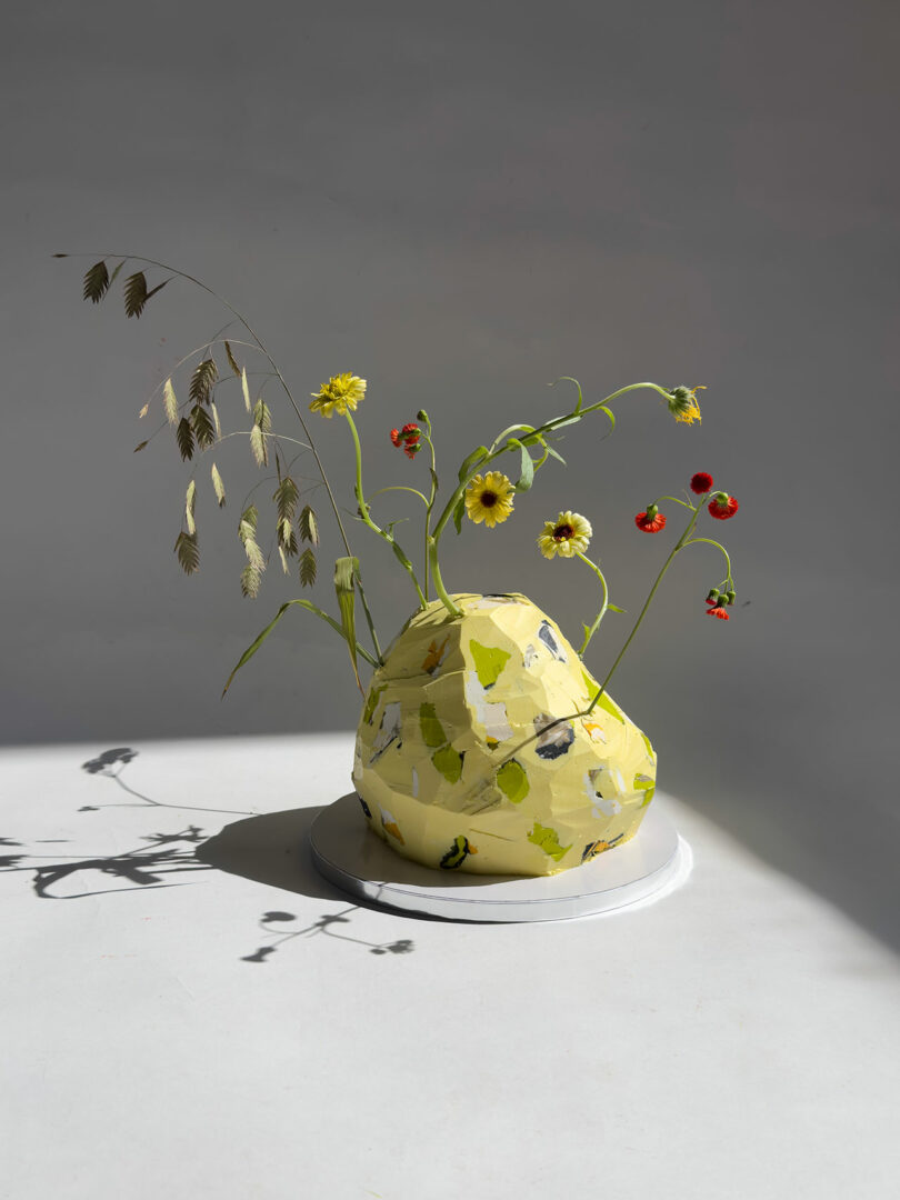 A yellow, irregularly shaped vase with a leaf pattern holds drooping wildflowers and grasses on a white surface, casting a shadow in bright, direct light.