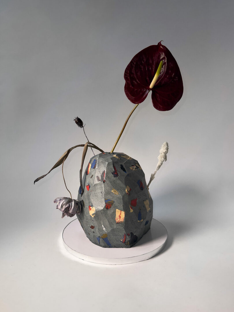 A speckled, geometric vase holds a red anthurium, dried flowers, and white stems, placed on a round white base against a neutral background.