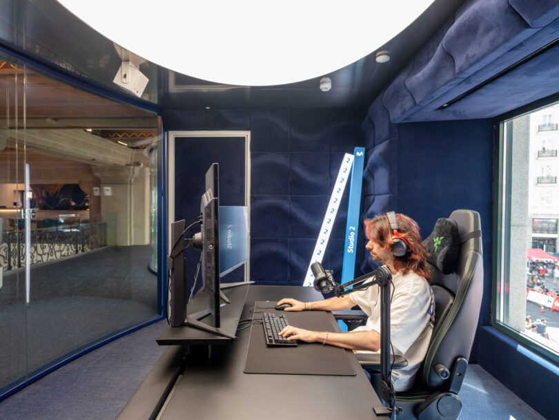 Person sitting in a soundproof room wearing headphones, using a computer with multiple screens and a microphone.