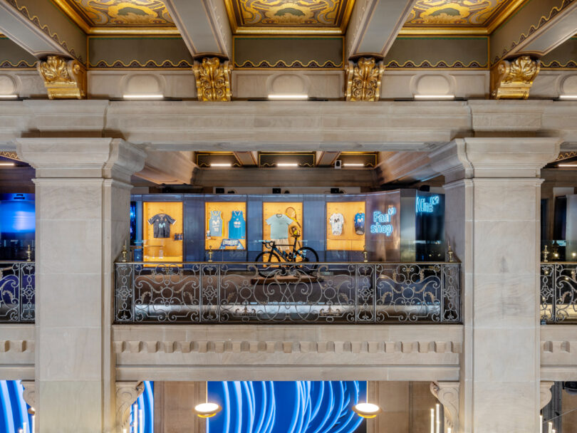 A balcony display in a store features sports jerseys, a bike, and a sign reading "Fan Shop," with decorative railings and ornate ceiling details.