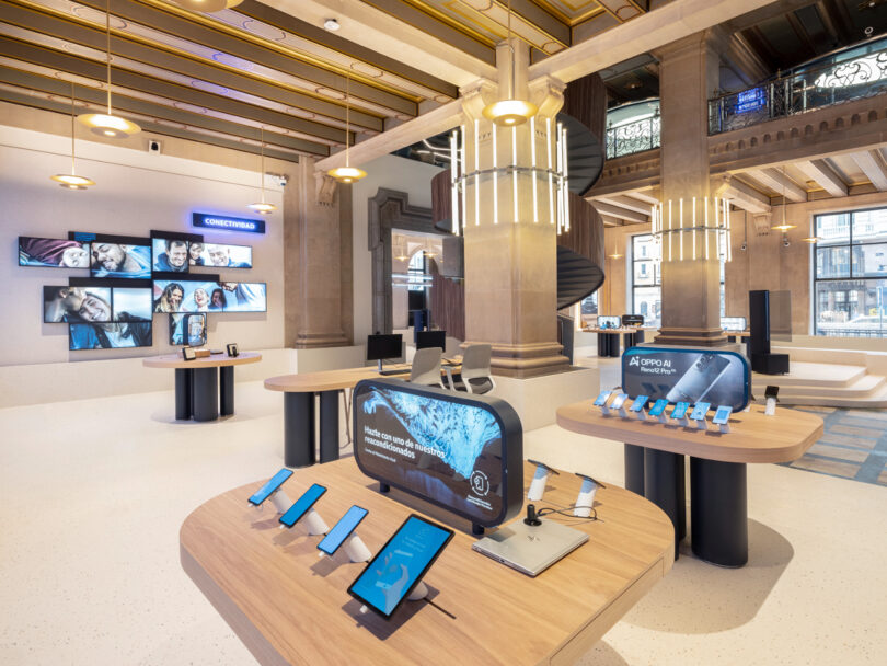 A modern electronics store interior with various smartphones and tablets displayed on wooden tables, digital screens on the walls, and a spiral staircase in the background.