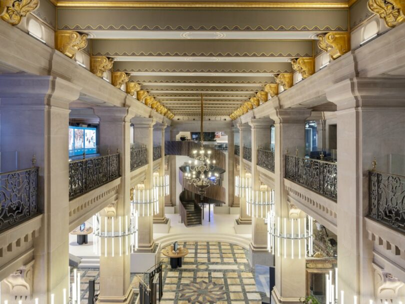 Interior of a grand hall with ornate columns, decorative ceiling, and a central chandelier. Marble floor features a geometric pattern. Dual staircases lead to an upper level with railing.