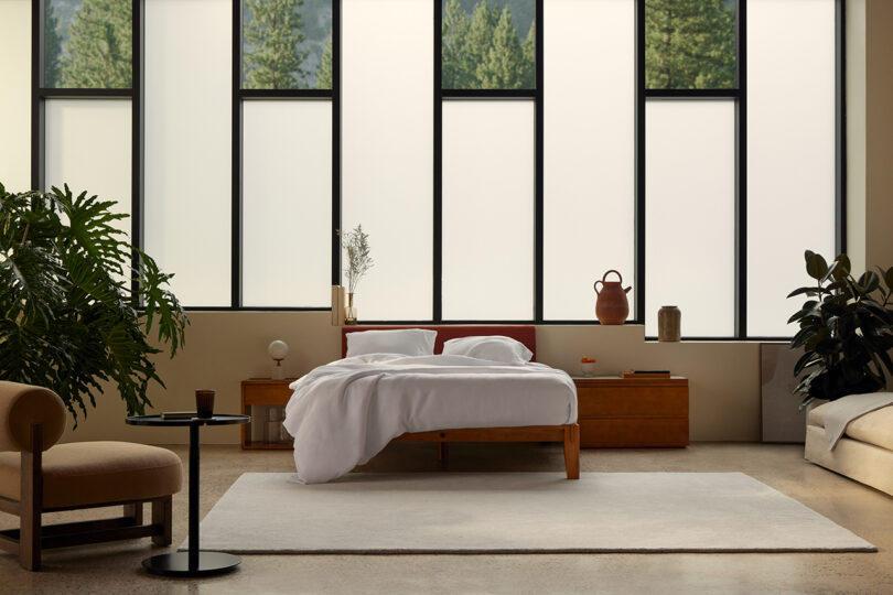 Modern bedroom with a wooden platform bed and headboard, white linens, side table with plant, and soft natural lighting.